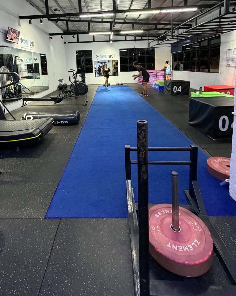 Blue turf track with weight plates and a sled, and members training in the distance